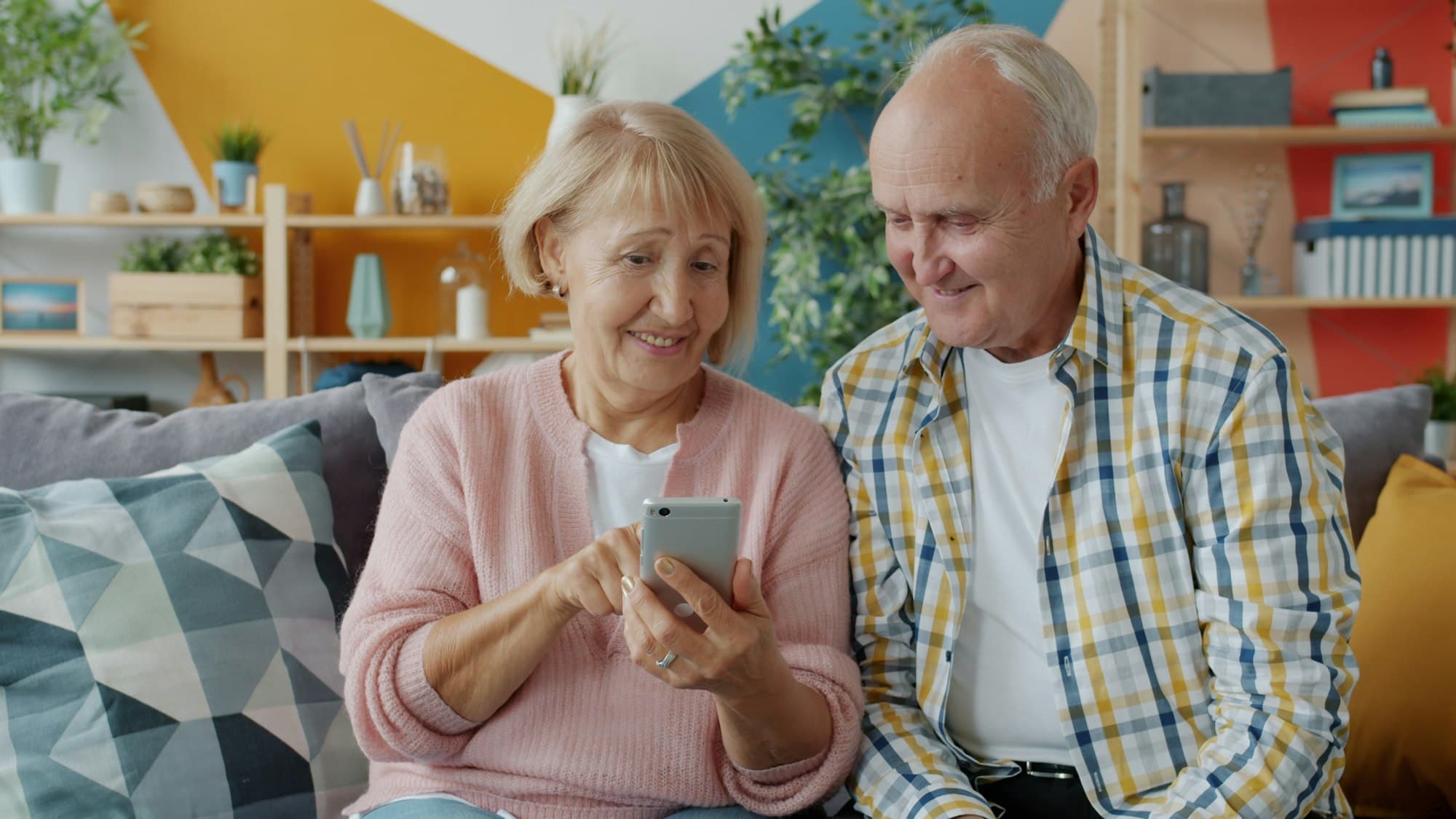 Casal de idosos sentado no sofá, sorrindo enquanto olha o celular junto