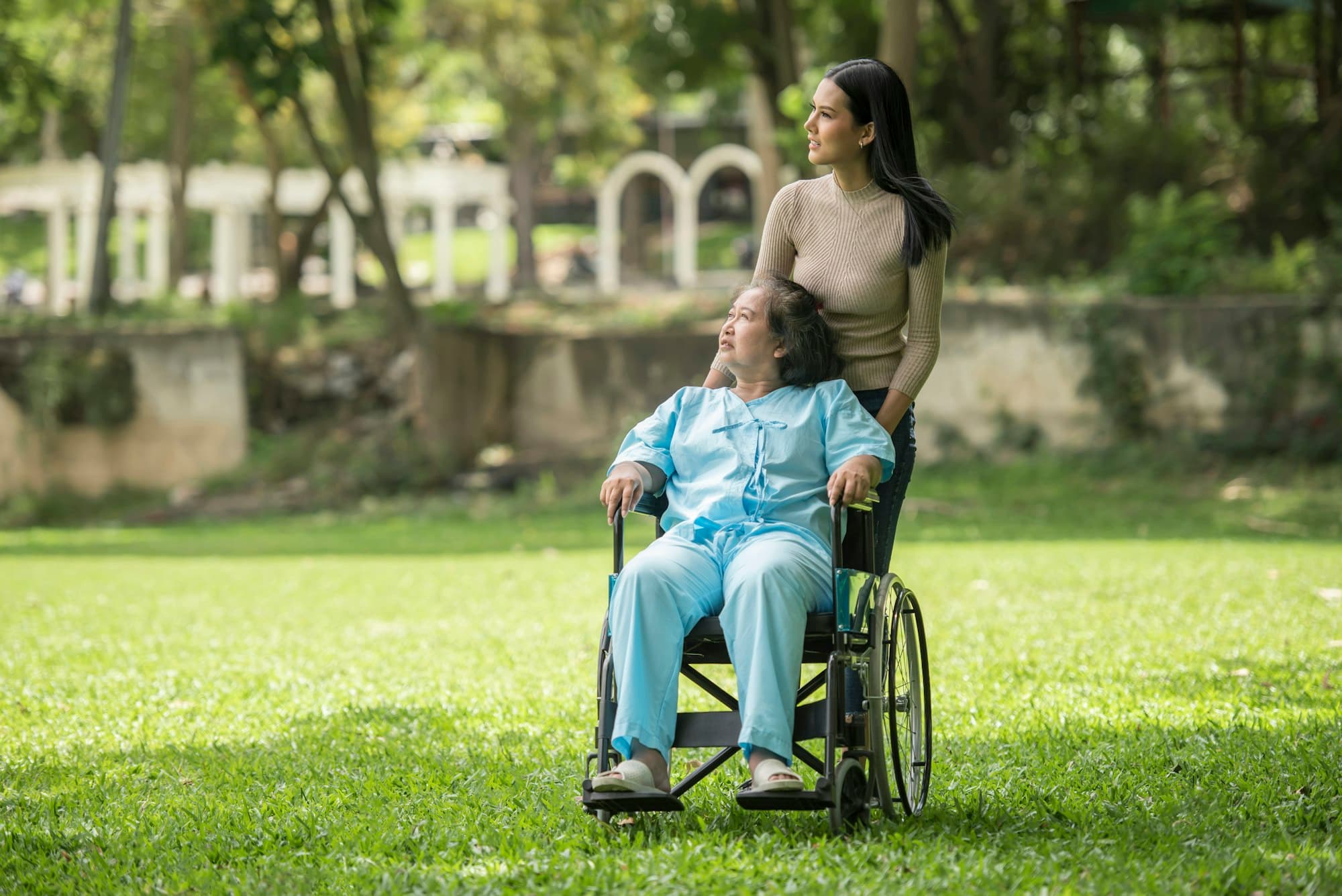 Moça empurra a cadeira de rodas de uma senhora num parque arborizado, em dia de sol